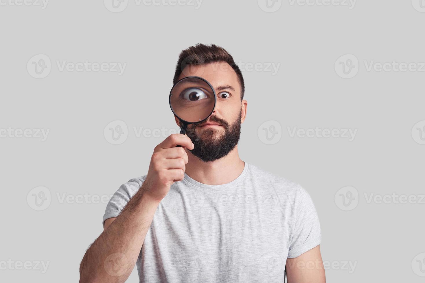 Modern detective. Playful young man applying magnifying glass while standing against grey background photo