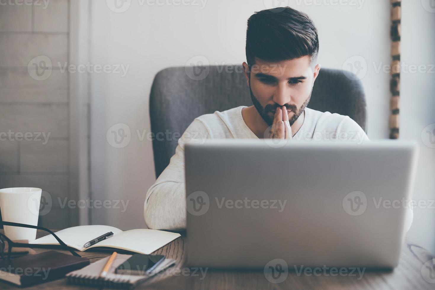 Praying for success. Front view of thoughtful young man holding hands on chin and looking at the laptop while sitting at his working place in office photo