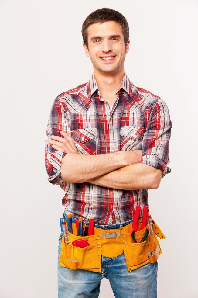 Cheerful handyman. Handsome young handyman with tool belt keeping arms crossed and smiling while standing against grey background photo