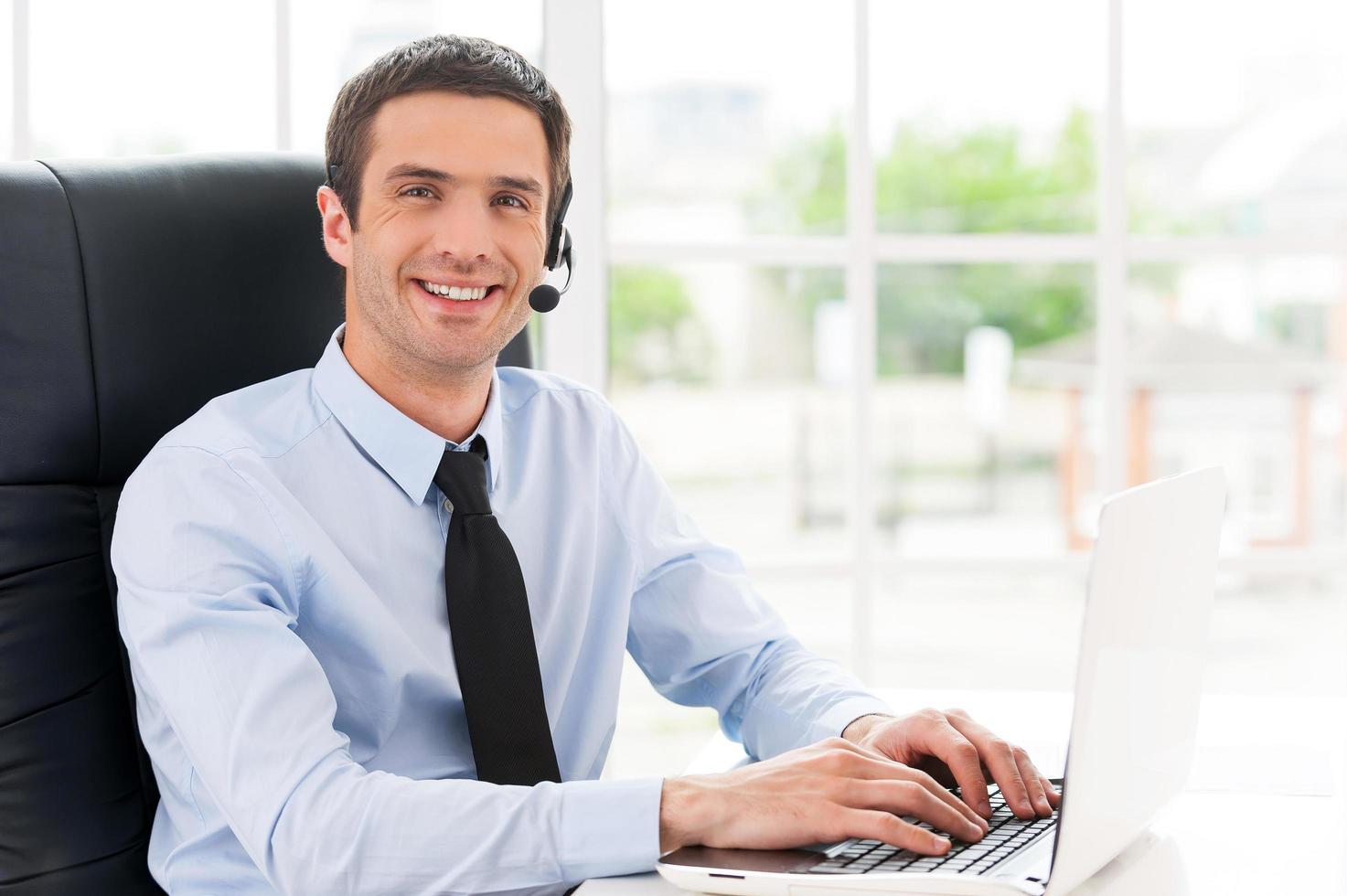 Cheerful male operator. Side view of cheerful young man in headset looking at camera and smiling while using laptop photo