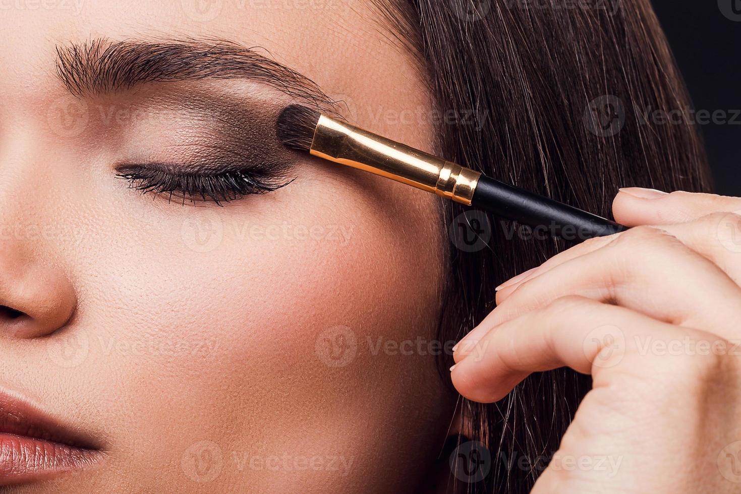 For perfect glance. Close-up of young woman applying eye shadow while standing against black background photo