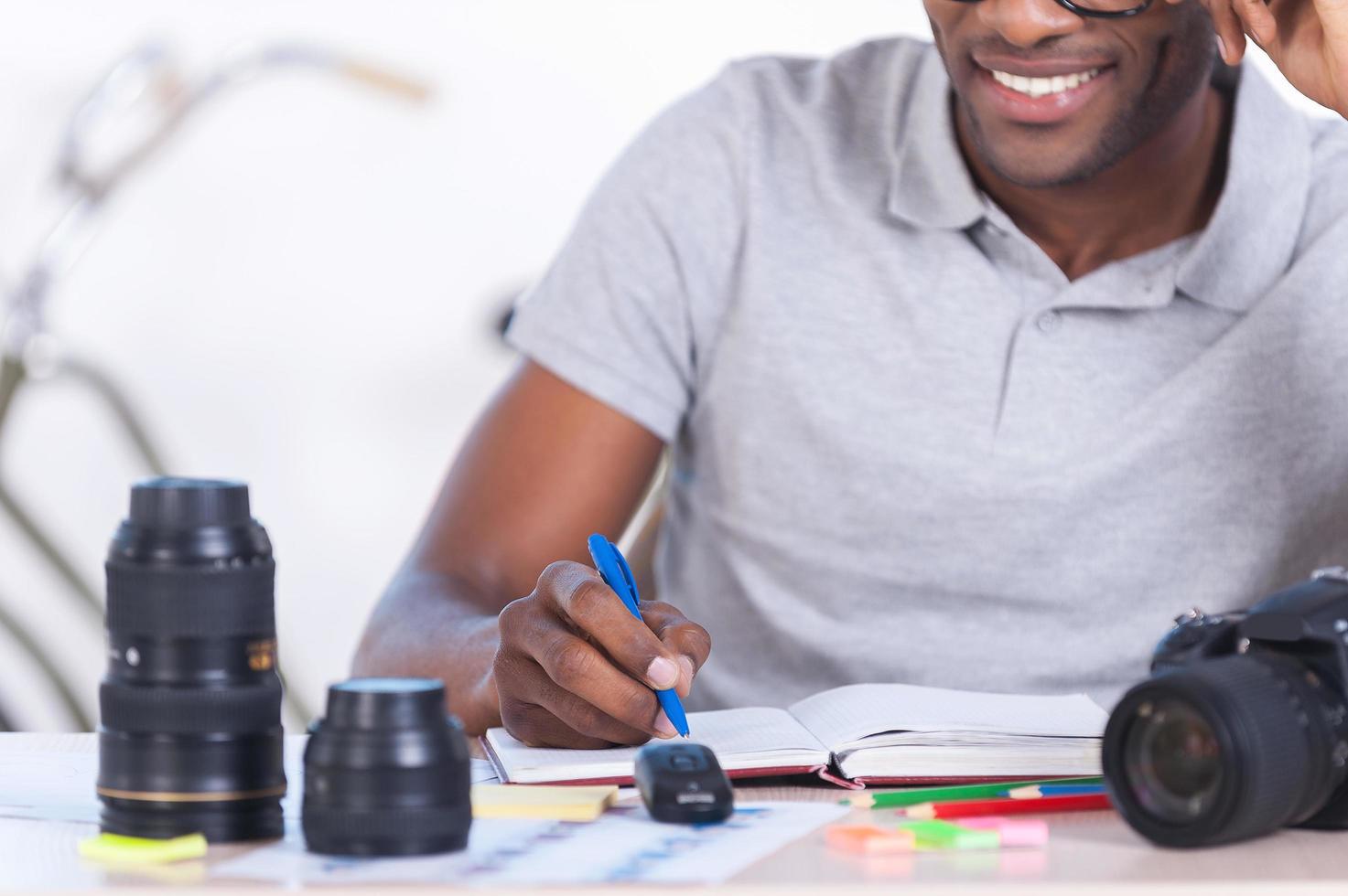 Working with pleasure. Cropped image of handsome young African man in casual wear sitting at his working place and writing something in his note pad photo