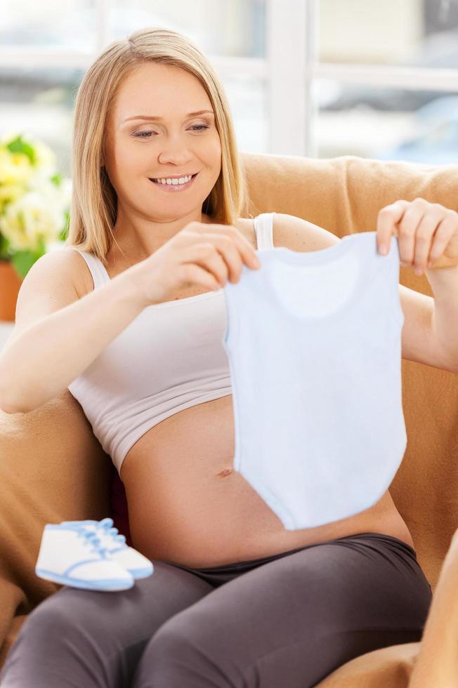 Pregnant woman with baby clothes. Beautiful pregnant woman sitting on the chair and showing baby clothing photo