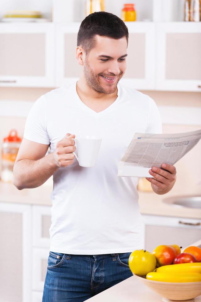 mañana sin preocupaciones. apuesto joven leyendo un periódico y sosteniendo una taza de café mientras está de pie en la cocina foto
