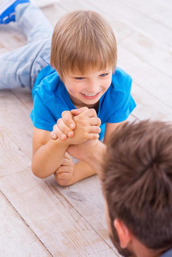 fuerte como su padre. vista superior de feliz padre e hijo compitiendo en la lucha de brazos mientras ambos yacen en el suelo de madera dura foto