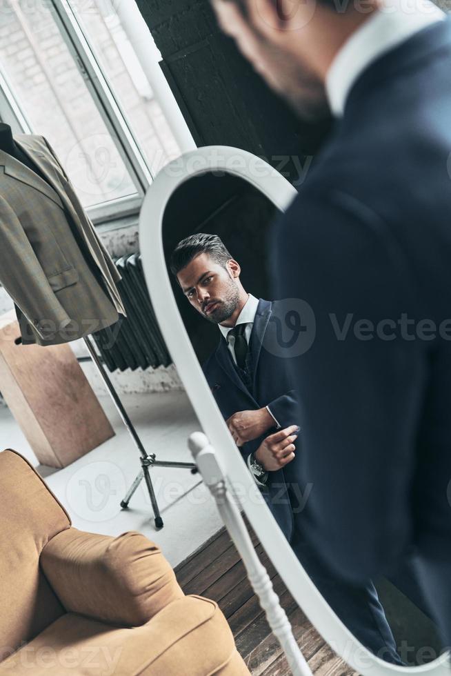 Confidence and charisma. Reflection of handsome young man in full suit adjusting his jacket while standing in front of the mirror indoors photo