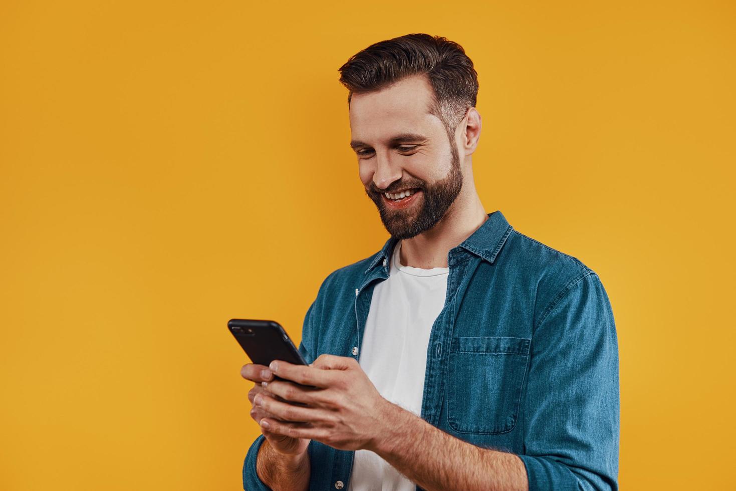 Charming young man in casual clothing using smart phone and smiling while standing against yellow background photo