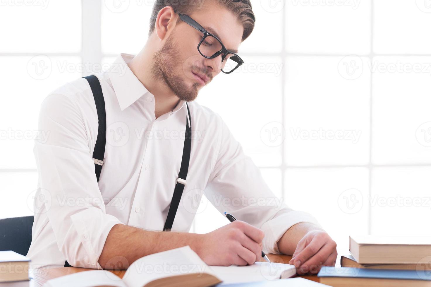 Author at work. Confident young man in shirt and suspenders writing something in note pad while sitting at his working place photo