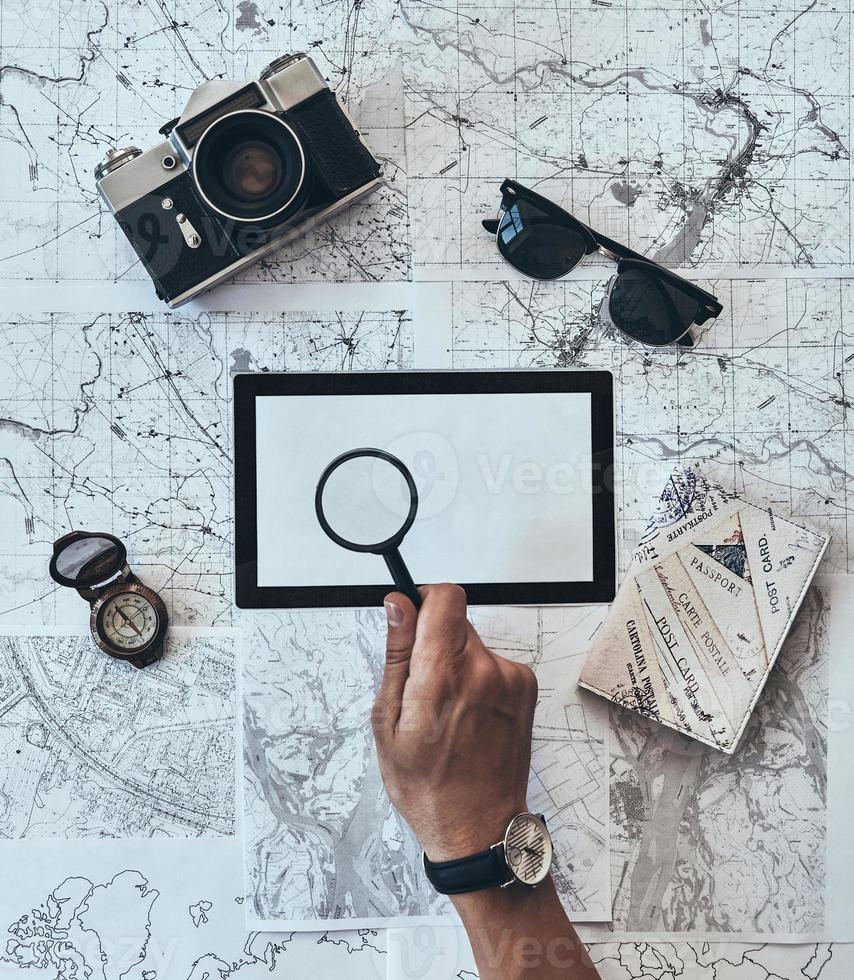 Trying to see everything. Close up top view of man using magnifying glass on copy space with sunglasses, photo camera, compass, passport lying on map around