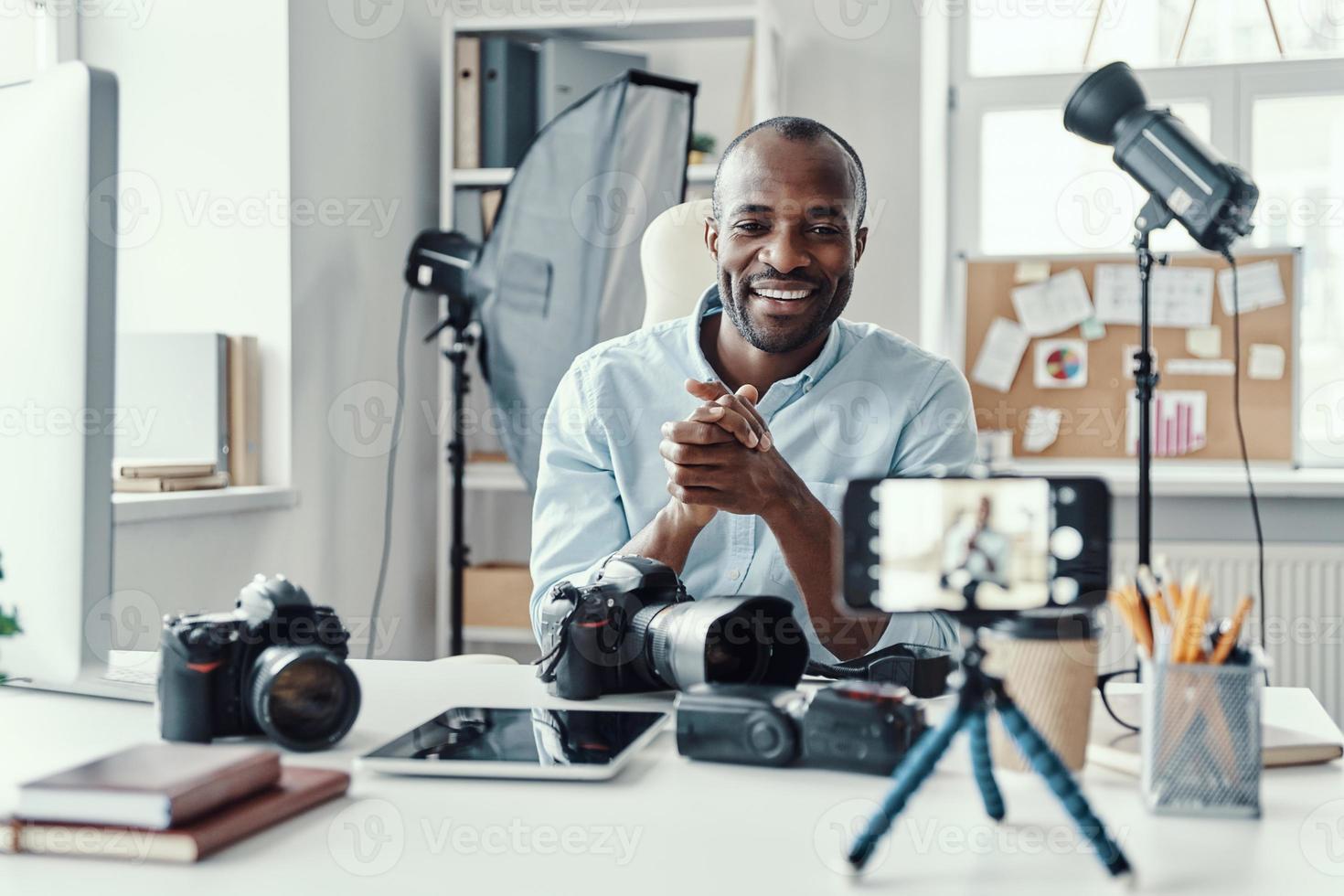 Charming young African man in shirt showing digital camera and telling something while making social media video photo
