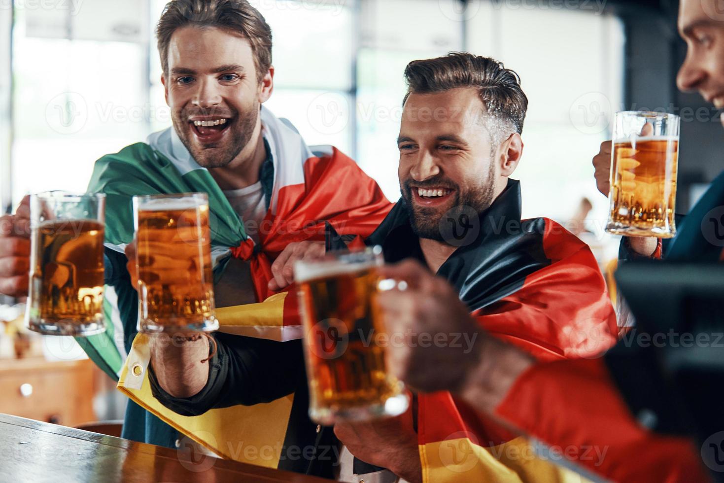 Happy young men covered in international flags toasting with beer while watching sport game in the pub photo
