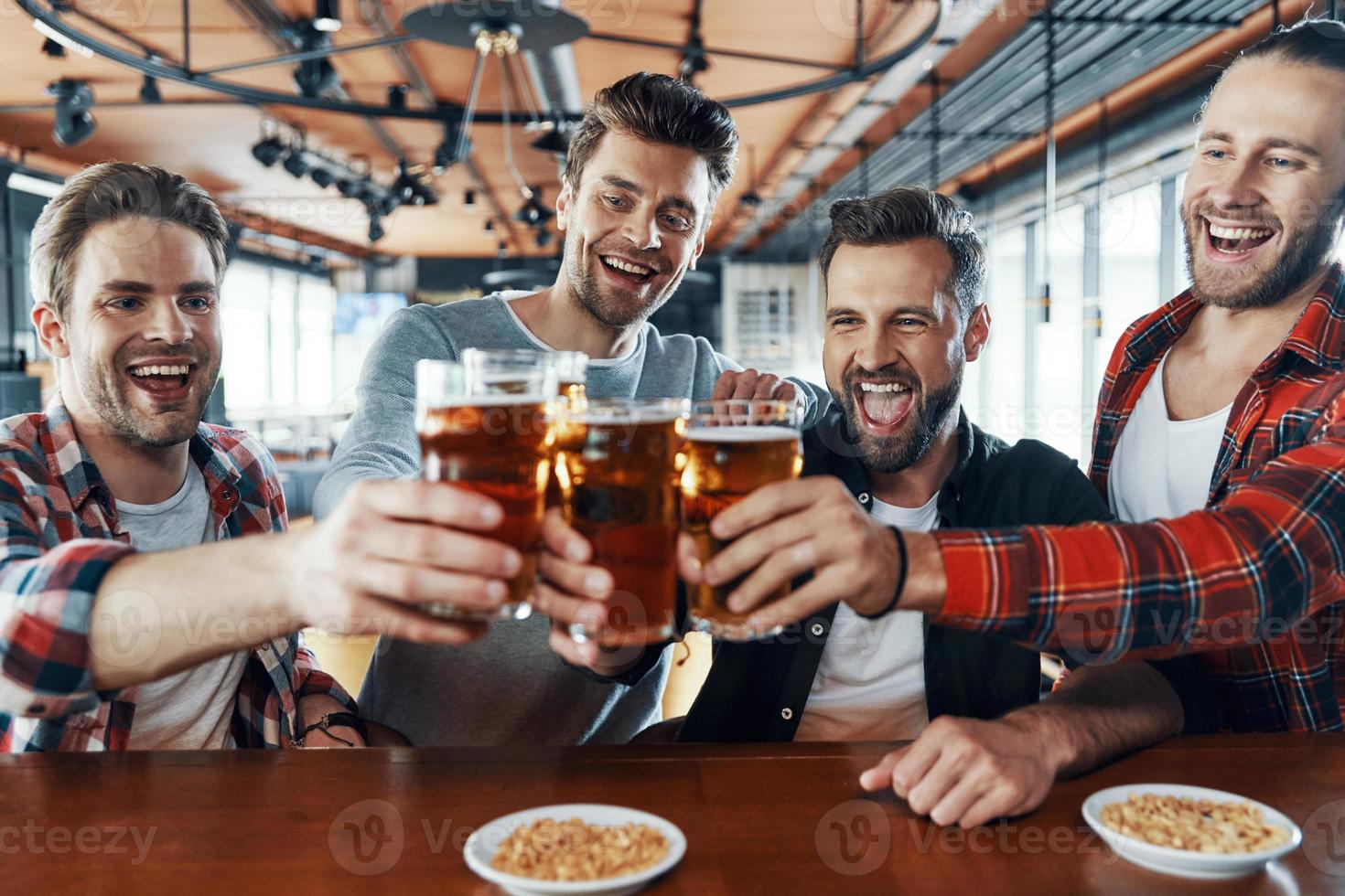 Happy young men in casual clothing toasting each other with beer and laughing while sitting in the pub photo