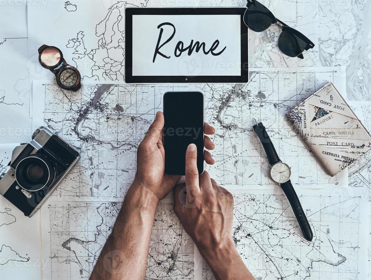 Rome. Close up top view of man using smart phone with sunglasses, photo camera, compass, watch and passport lying on map around