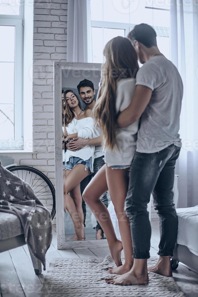 Perfect couple. Full length rear view of young people bonding and smiling while looking in the mirror at home photo