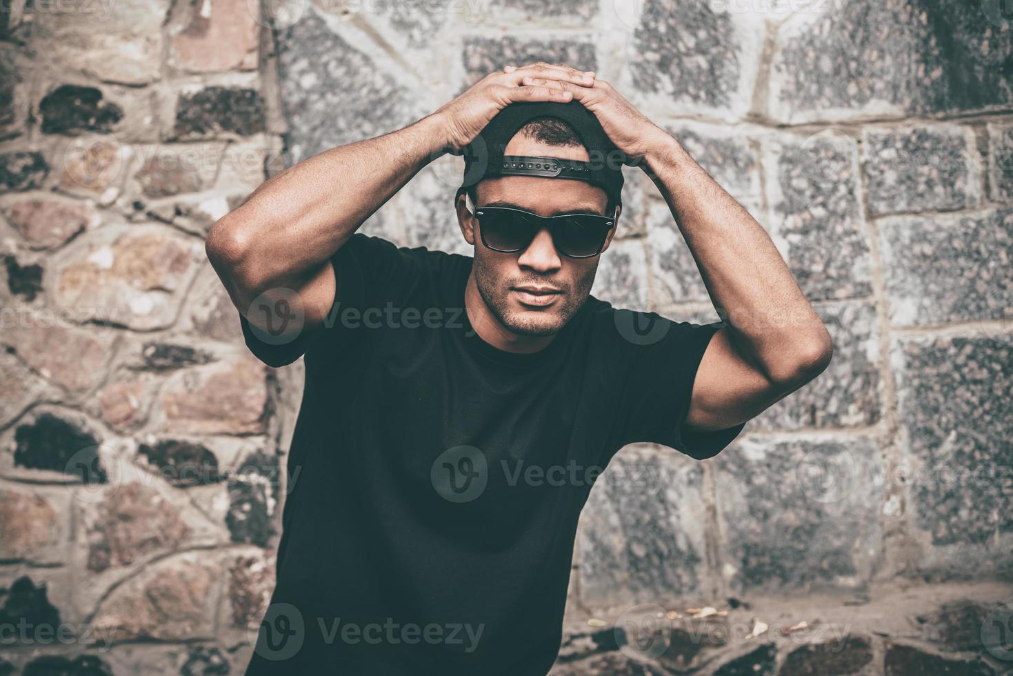 Young and cool. Handsome young African man in casual clothes holding hands on cap and looking at camera while standing against the stoned wall outdoors photo