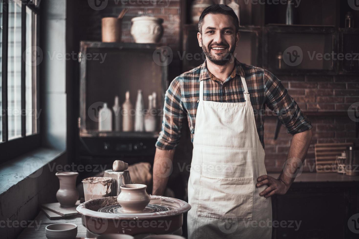 Cheerful potter. Confident young man looking at camera and smiling while standing in pottery photo