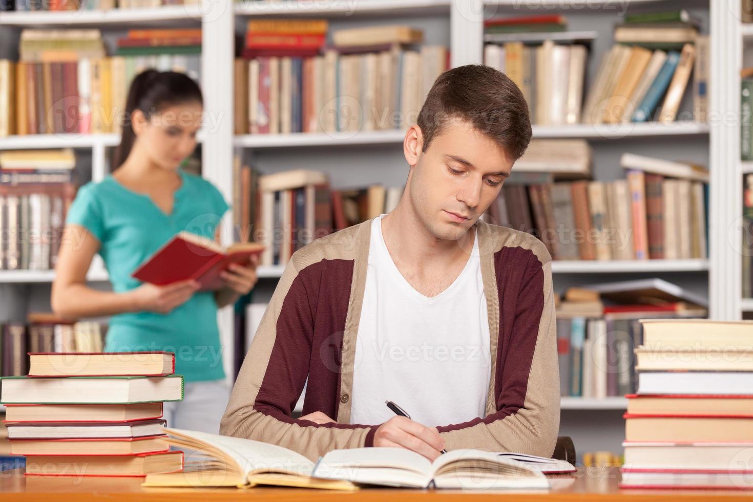 Preparing to the exams. Thoughtful young man sitting at the library desk and writing something in his note pad while young woman reading a book on the background photo