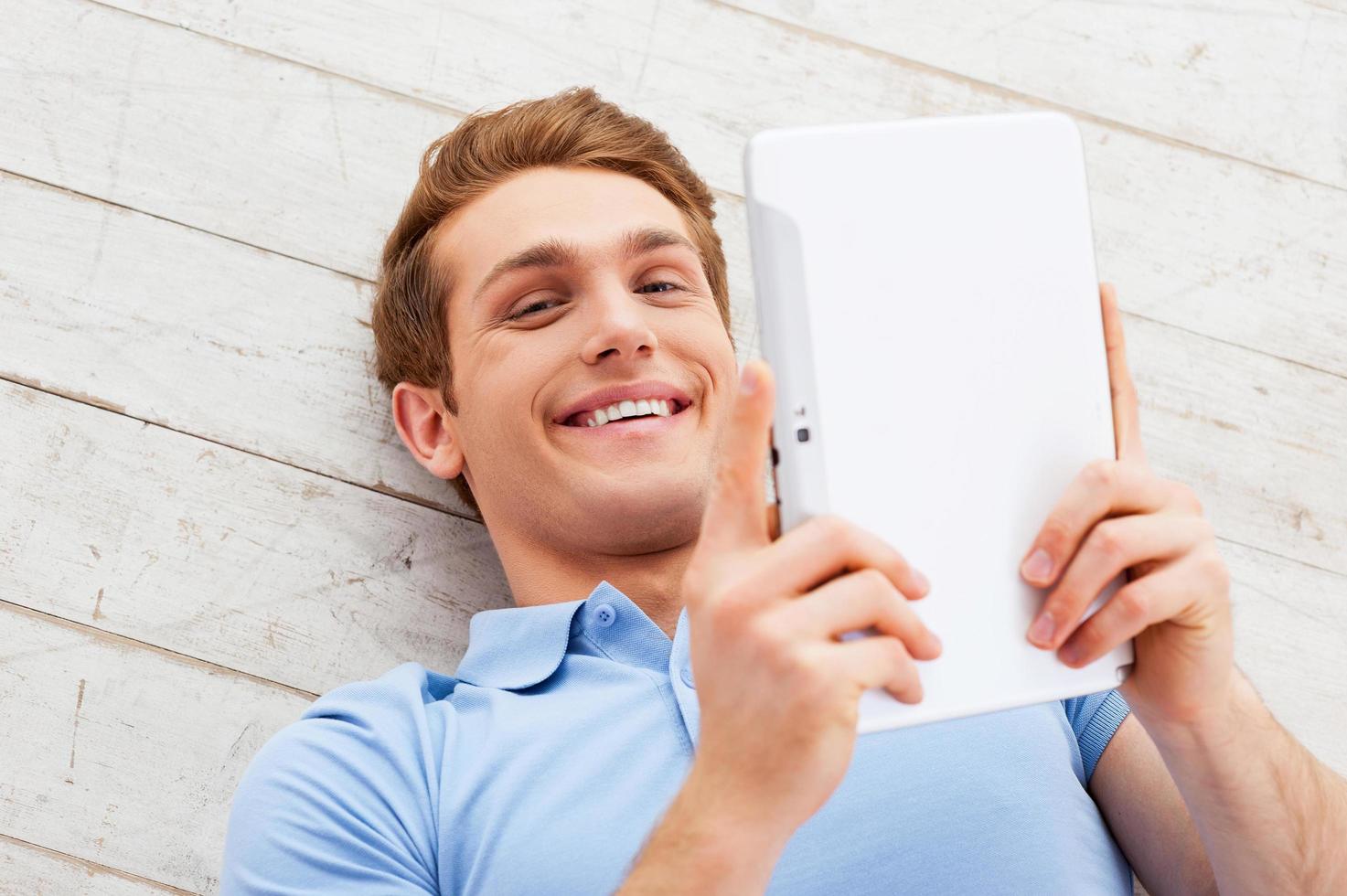 Examining his new gadget. Top view of happy young man lying on the floor and holding digital tablet photo