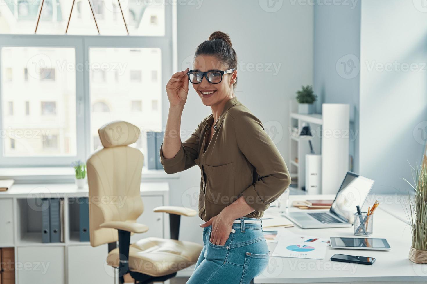 hermosa mujer joven con ropa informal elegante sonriendo y ajustando los anteojos mientras está pie en la oficina 13540919 Foto de stock en Vecteezy