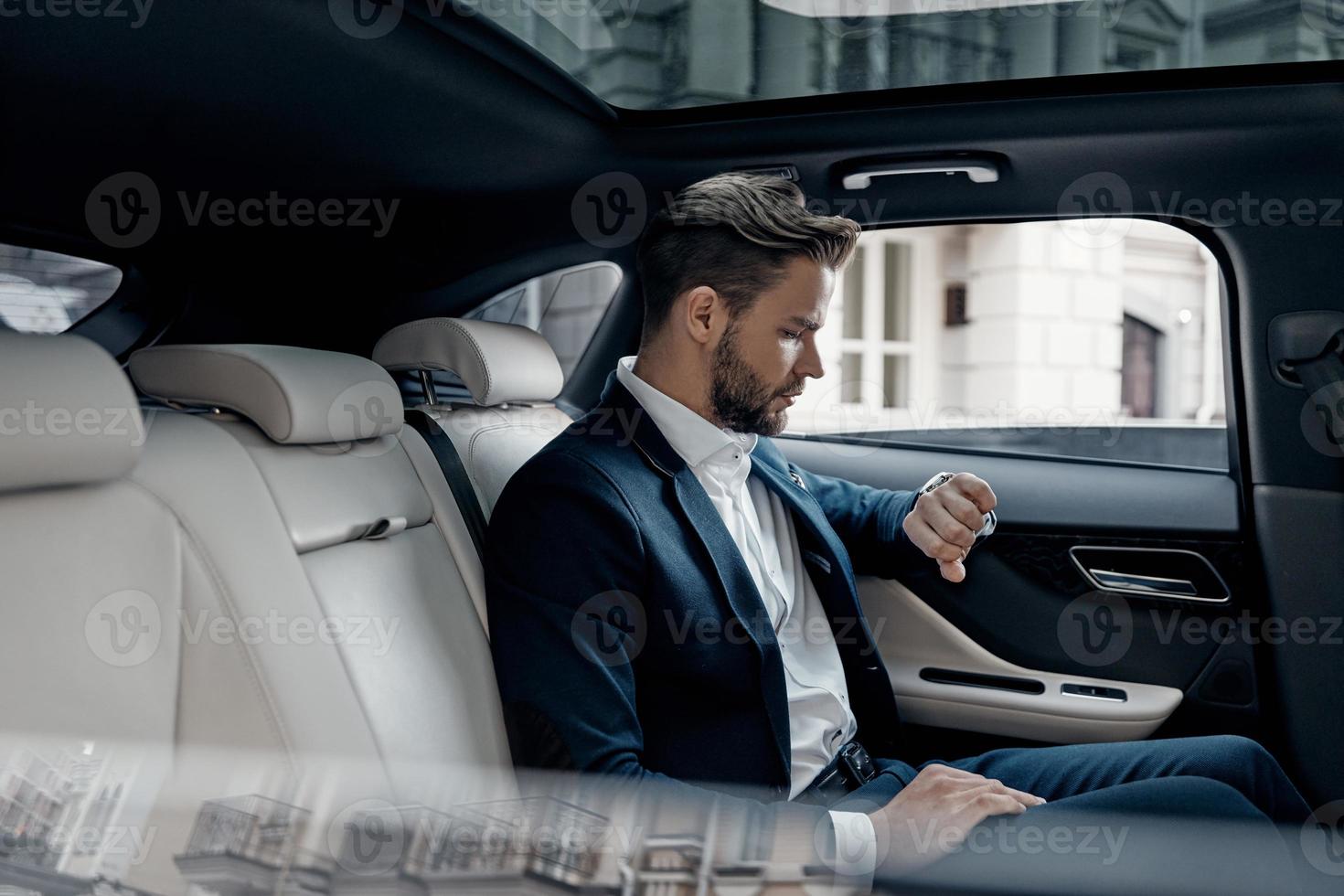 Just in time. Handsome young man in full suit looking at his watch while sitting in the car photo