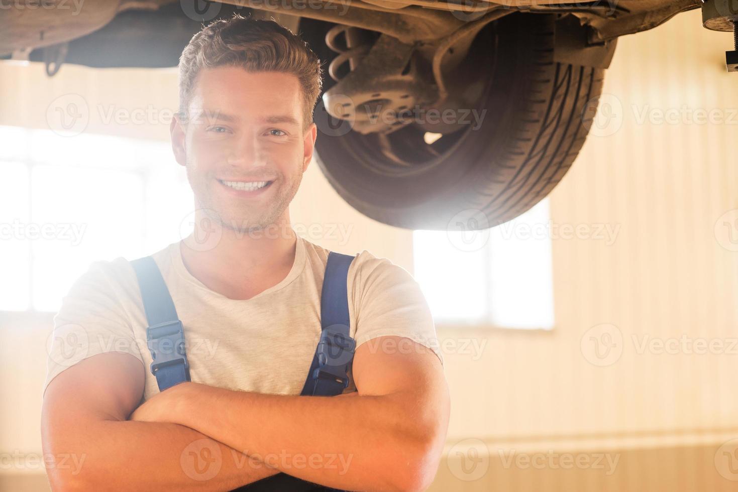Confident professional. Handsome young man in uniform keeping arms crossed and smiling while standing underneath a car in workshop photo