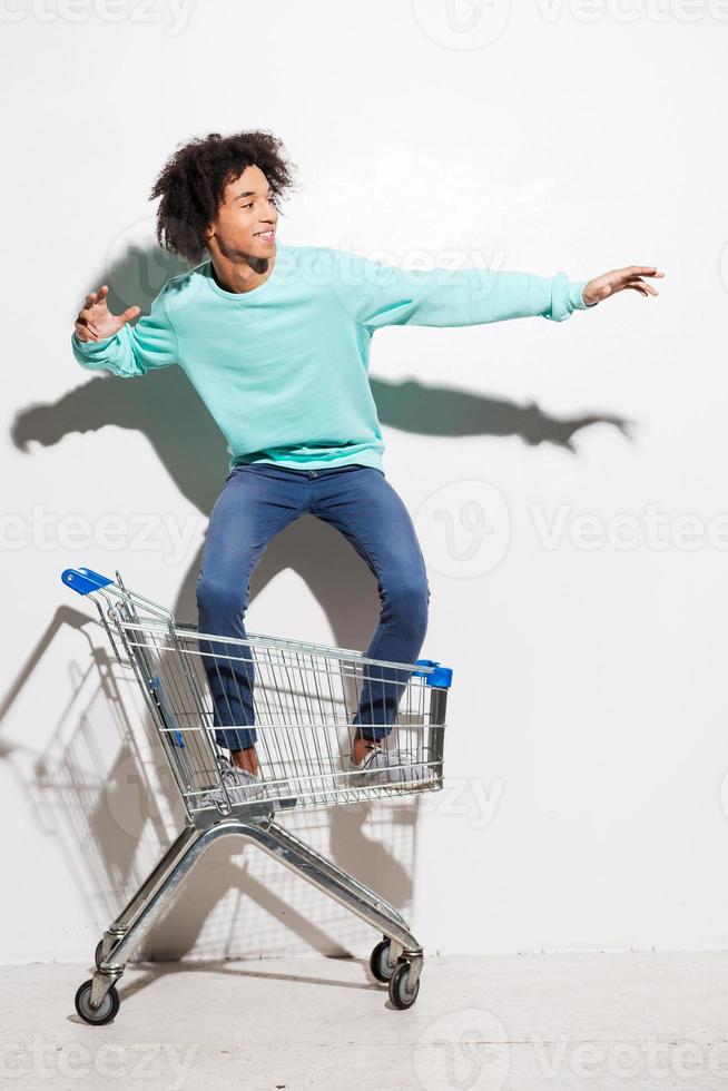 montando un carrito de compras. un joven africano juguetón montando en un carrito de compras contra un fondo gris foto