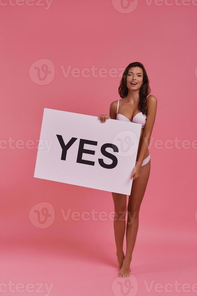 Quick answer.  Full length of attractive young woman holding a poster and smiling while standing on tiptoe against pink background photo