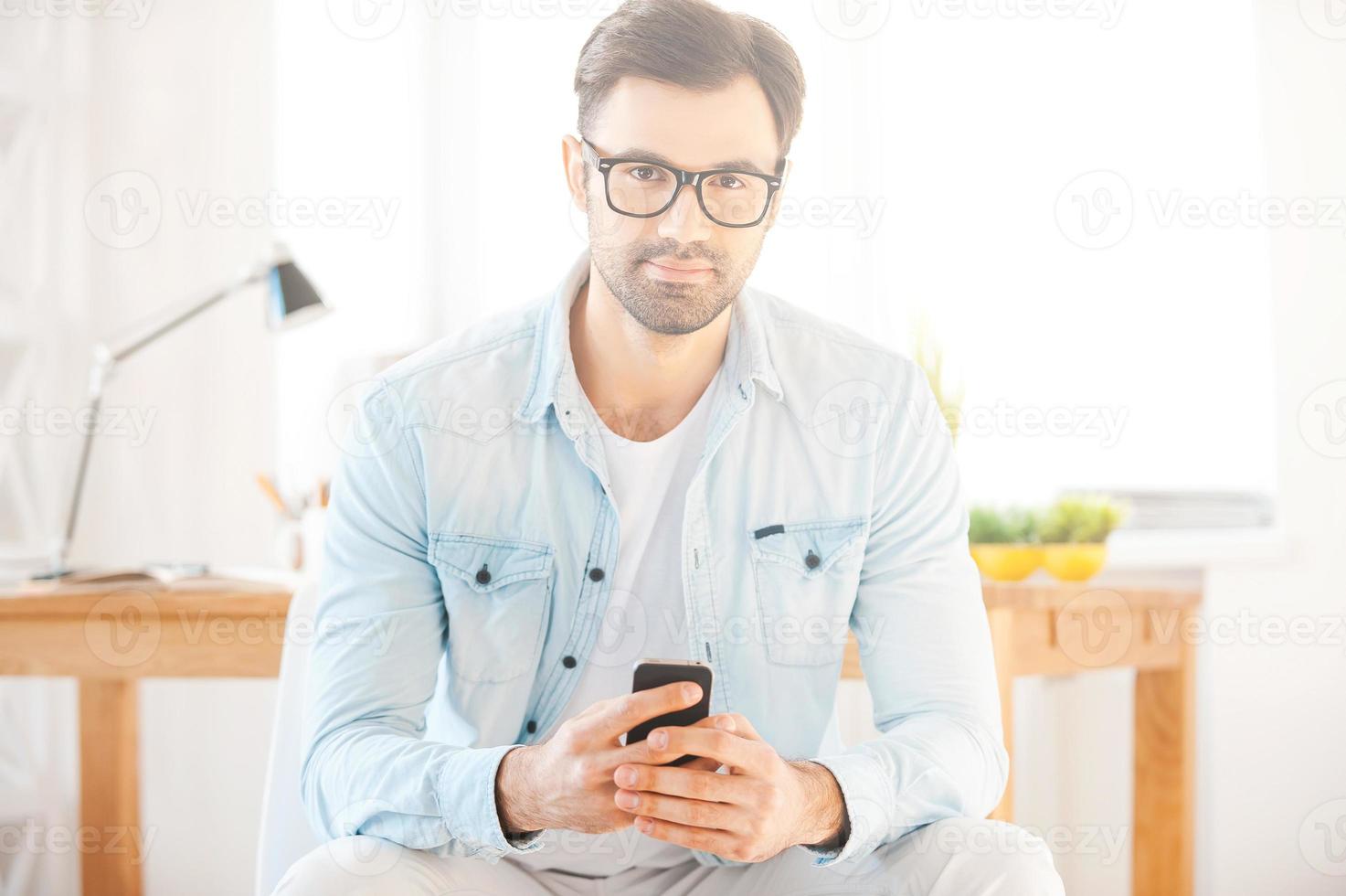 My work is my life. Handsome young man in shirt and eyewear holding mobile phone and looking at camera while sitting in front of his working place photo