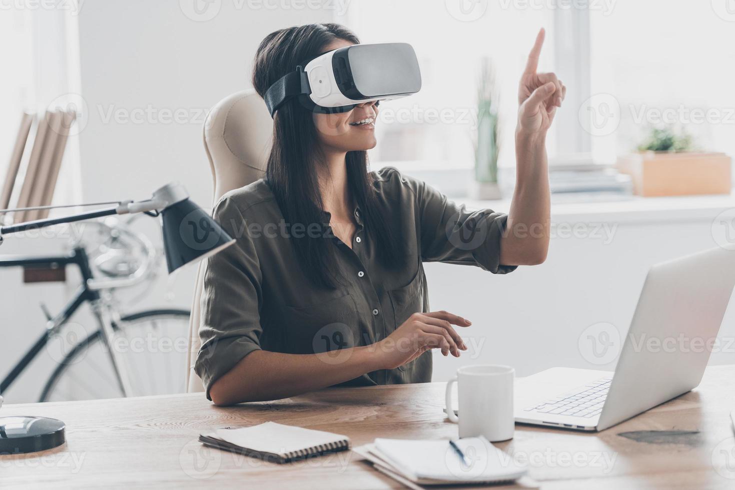 usando tecnologías vr. joven segura de sí misma con auriculares de realidad virtual apuntando al aire mientras se sienta en su lugar de trabajo en la oficina foto