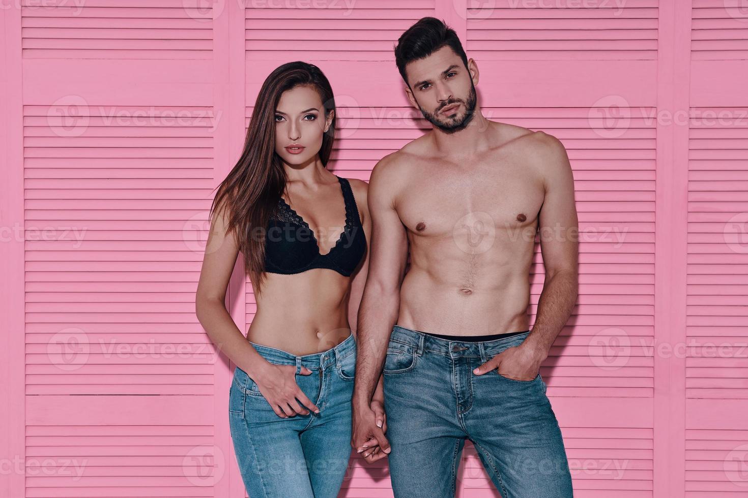 Made to love each other. Beautiful young couple holding hands and looking at camera while standing against pink background photo