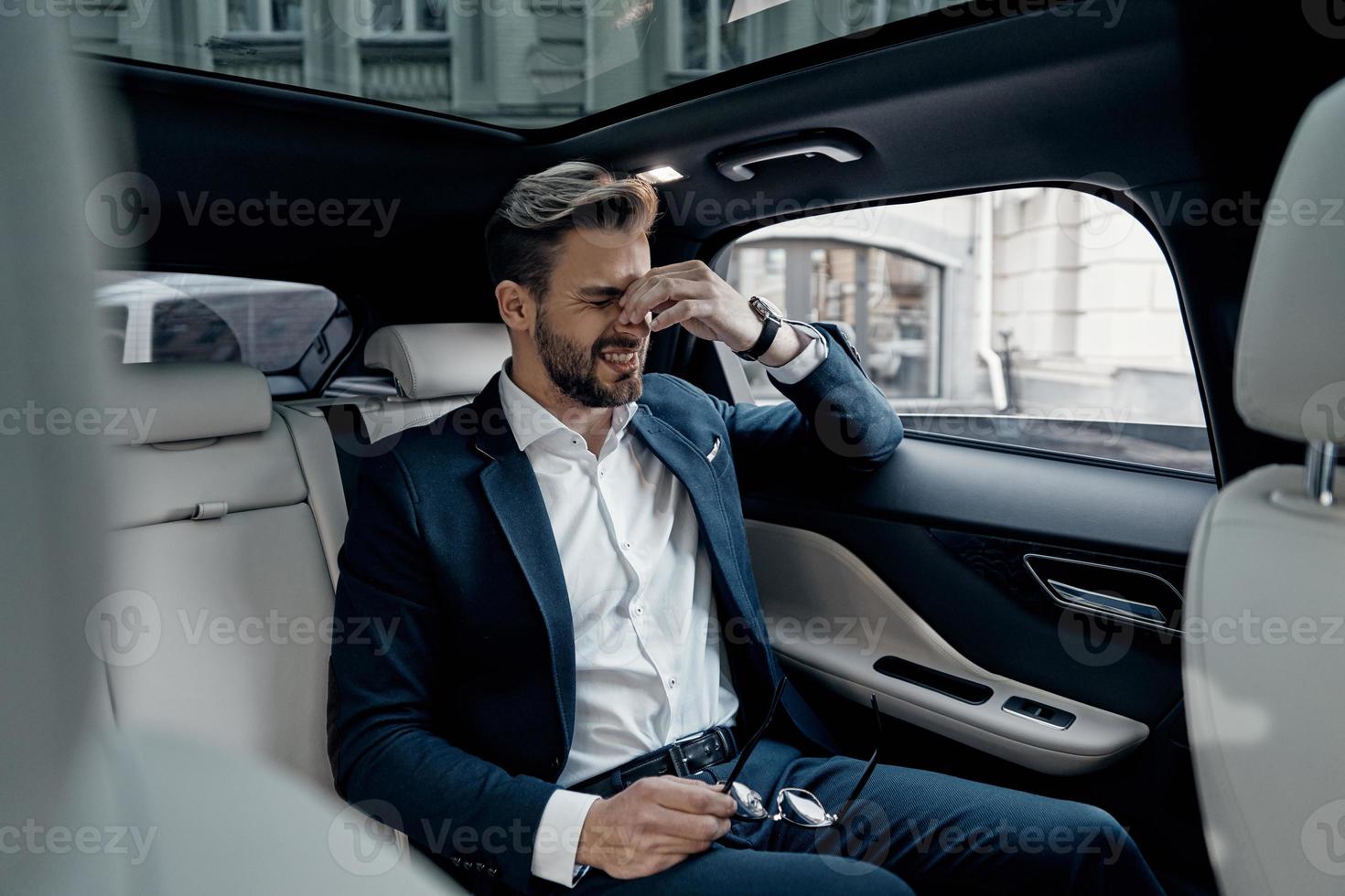 teniendo dolor de cabeza. joven frustrado con ropa formal masajeando la nariz y manteniendo los ojos cerrados mientras se sienta en el auto foto