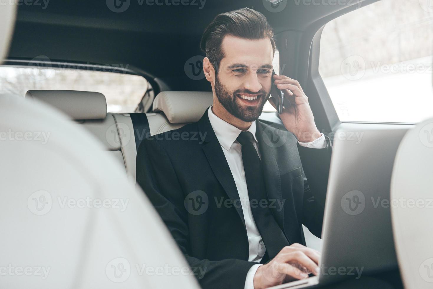 centrándose en el trabajo. apuesto joven hombre de negocios trabajando en su computadora portátil y hablando por teléfono mientras está sentado en el asiento trasero del camión foto