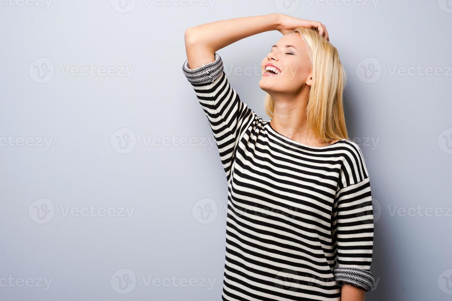 divertido y coqueto. hermosa joven mujer de cabello rubio manteniendo la mano en el cabello y sonriendo mientras está de pie contra el fondo gris foto