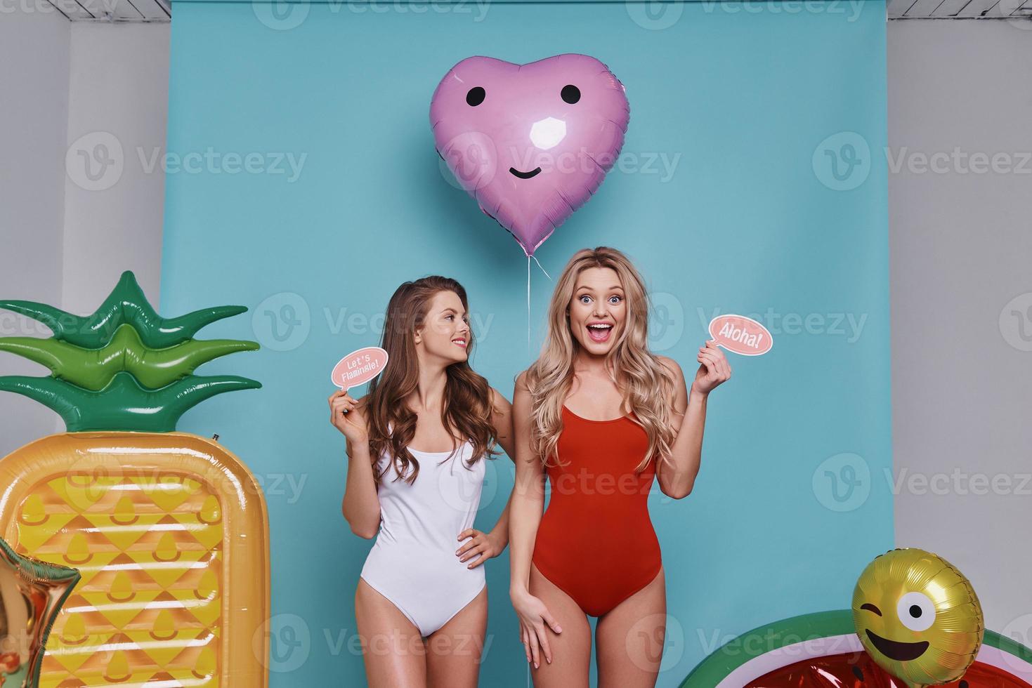Carefree and happy.  Two playful young women in swimwear looking at camera and smiling while standing against blue background photo