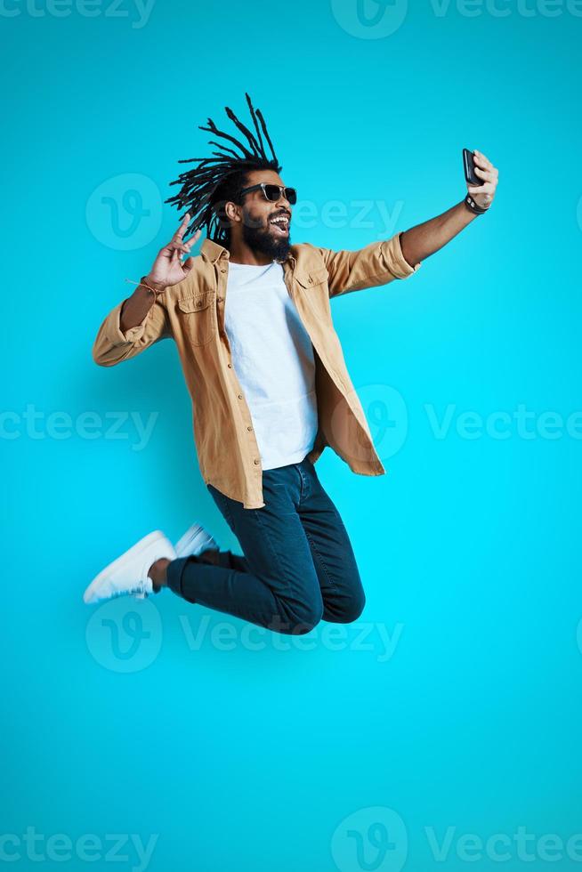 toda la longitud de un joven africano juguetón con ropa informal tomándose selfie y sonriendo mientras flotaba contra el fondo azul foto