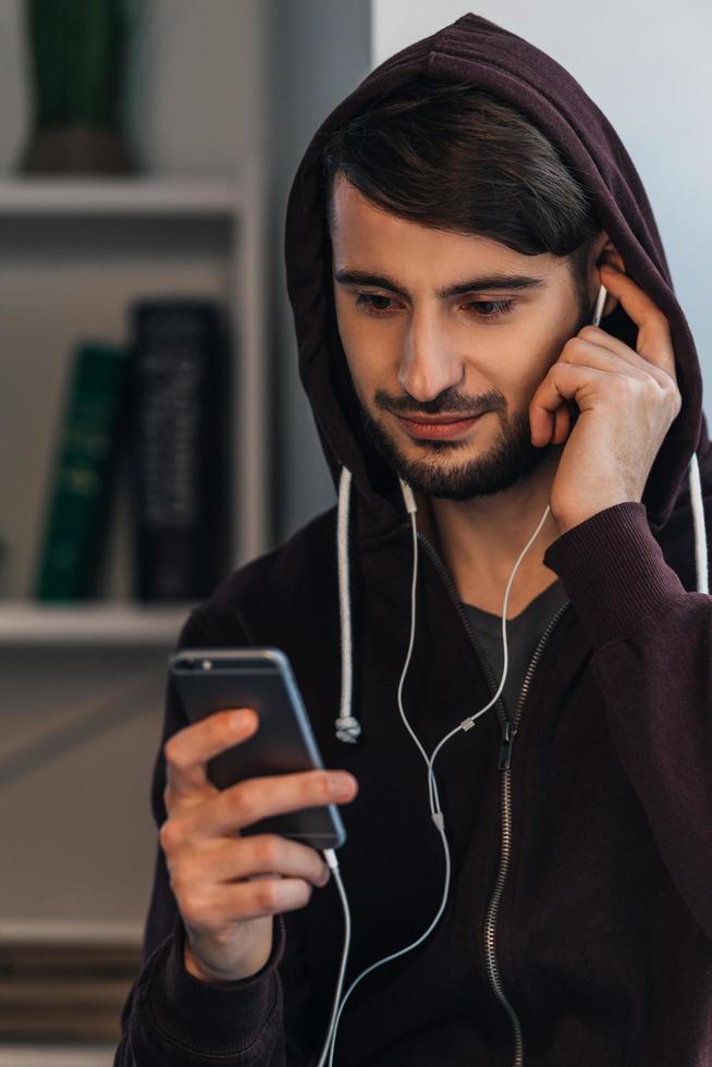 Next song. Pensive young man in hooded shirt using his smartphone while standing at home photo