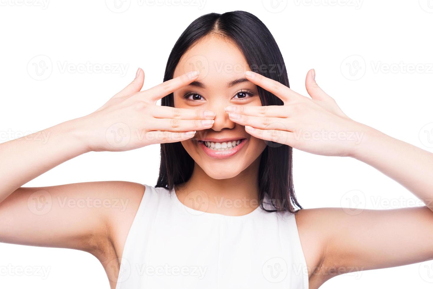 Playful beauty. Beautiful young Asian woman in pretty dress looking at camera and covering her face by hands while standing against white background photo