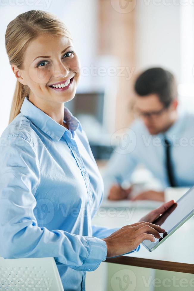 Working hard to become successful. Smiling young businesswoman in formalwear holding digital tablet and looking at camera while her male colleague working in the background photo