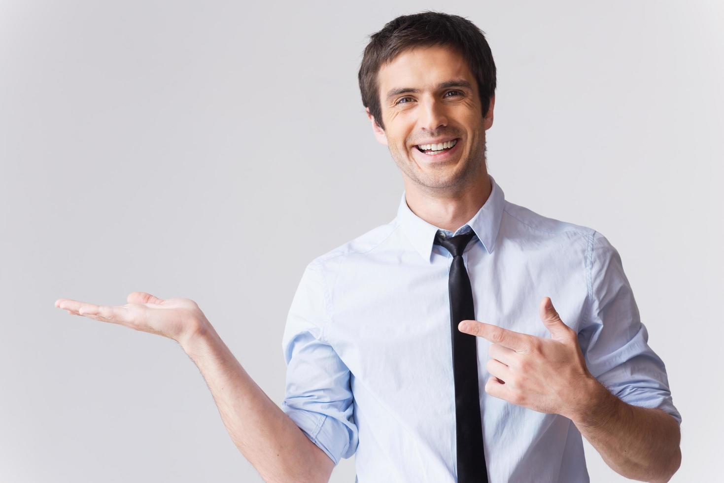 Businessman holding copy space. Happy young man in shirt and tie holding copy space on his hand and smiling while standing against grey background photo