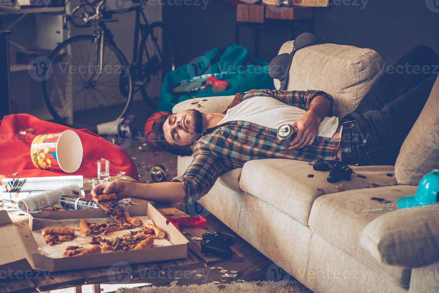 bebió demasiada cerveza. joven apuesto se desmayó en el sofá con una rebanada de pizza y una lata de cerveza en la mano en una habitación desordenada después de la fiesta foto