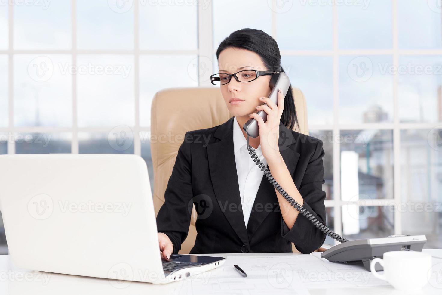 Businesswoman at work. Confident young business woman talking on the phone and using computer while sitting at her working place photo