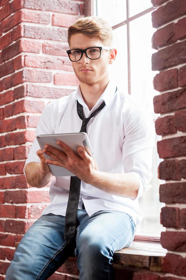 Handsome with digital tablet. Handsome young man in shirt and tie working on digital tablet and looking at camera while sitting at the window sill photo