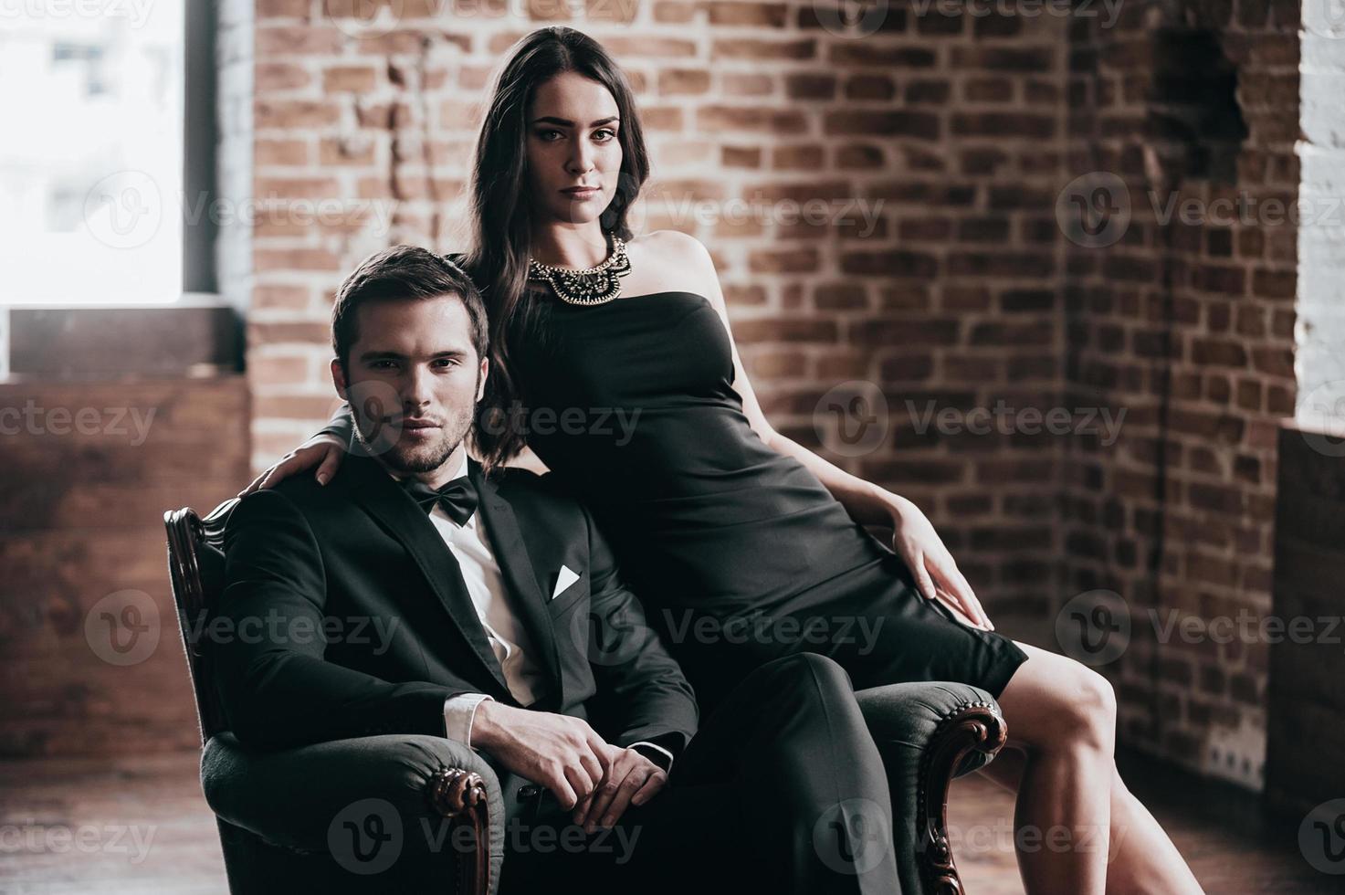 Perfect couple. Beautiful young couple in formalwear looking at camera while sitting in a chair in loft interior photo