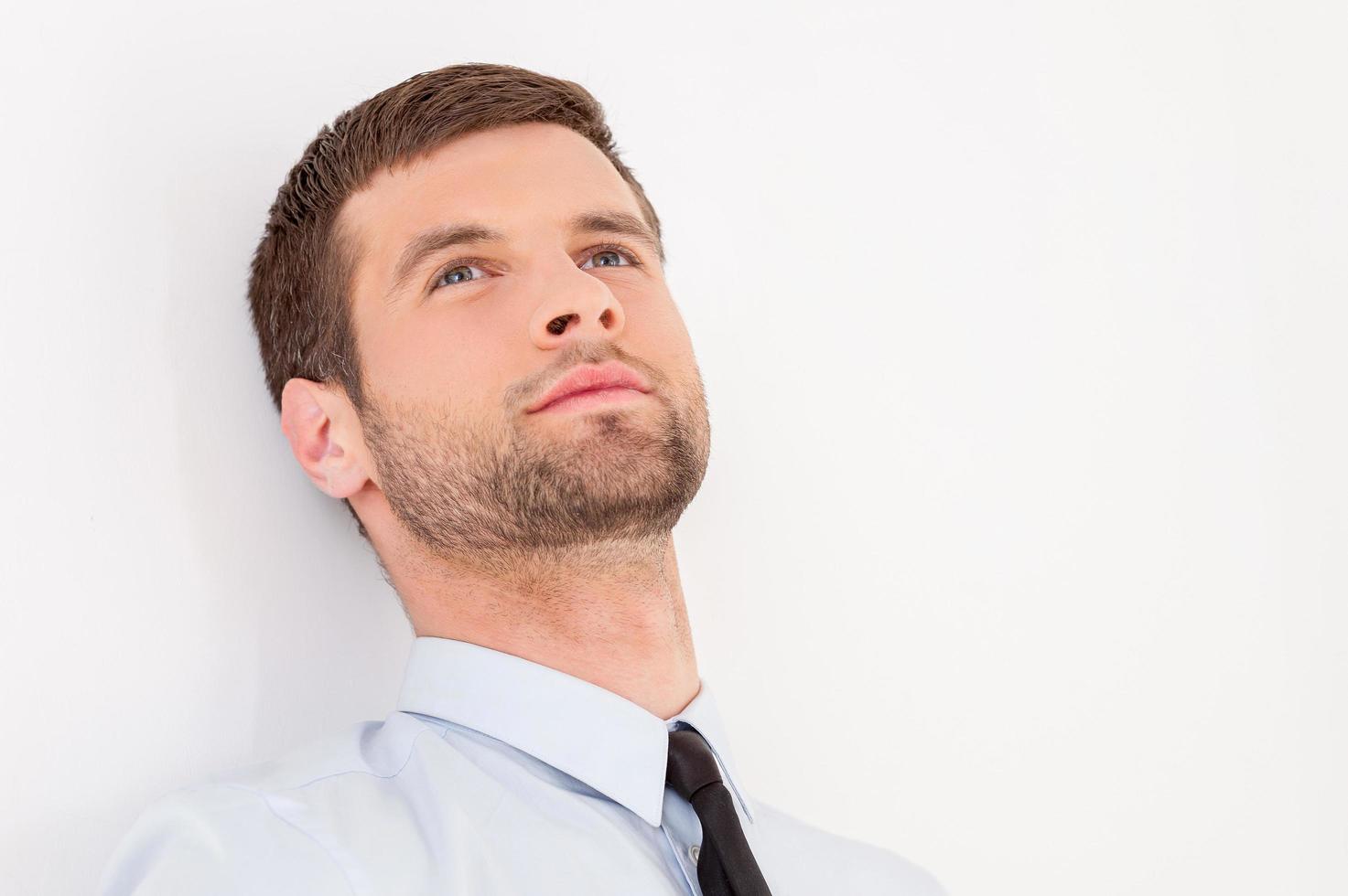 pensando en oportunidades de negocio. apuesto joven con camisa y corbata mirando hacia otro lado y sonriendo mientras se apoya en la pared foto