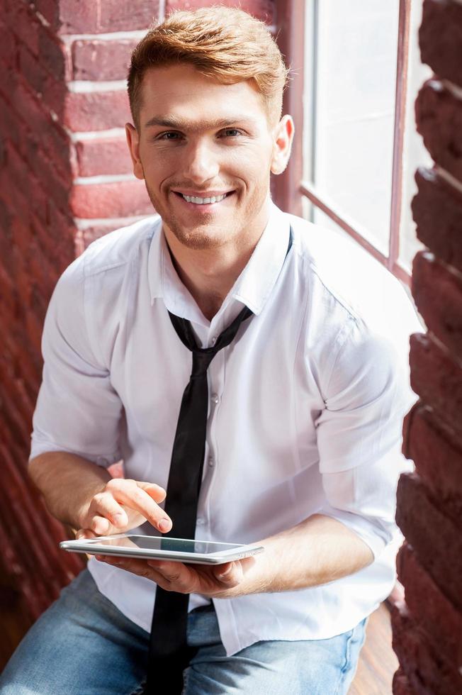 Testing his brand new tablet. Top view of handsome young man in shirt and tie working on digital tablet and smiling while sitting at the window sill photo