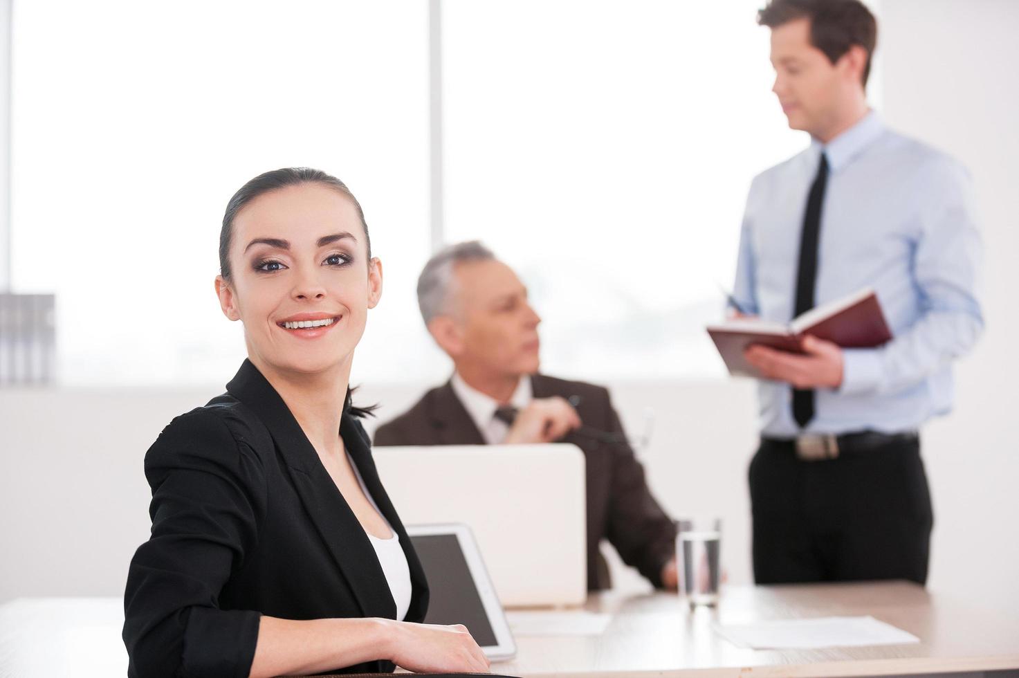 Happy to be a part of team. Attractive young woman in formalwear looking over shoulder and smiling while sitting at the table with two men talking on background photo
