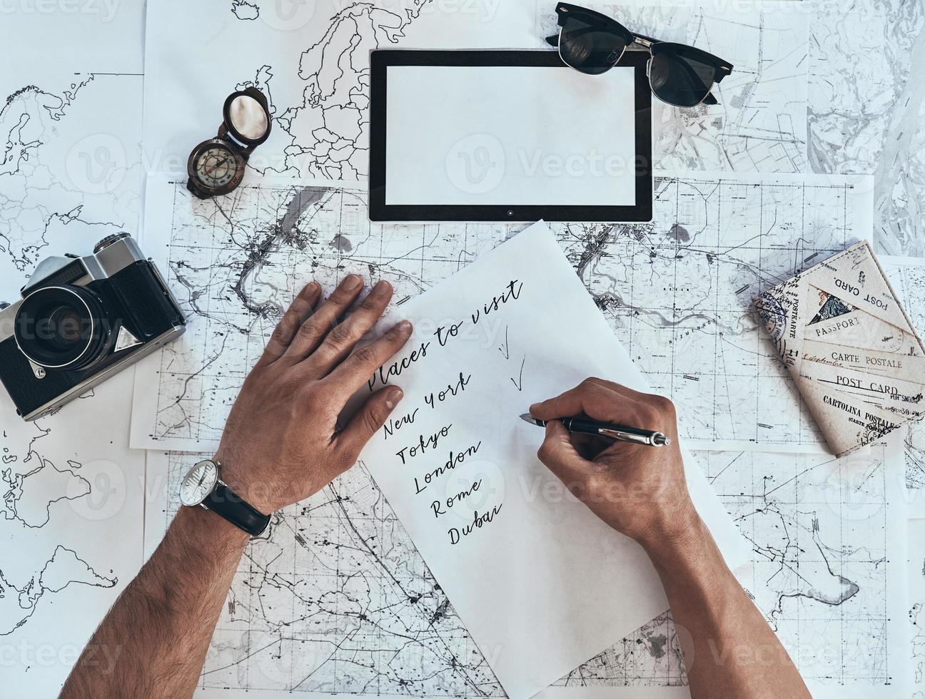 Making a list. Close up top view of man writing down checklist with sunglasses, photo camera, compass, passport lying on map around