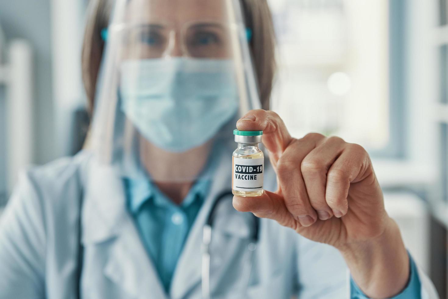 Mature female doctor in white lab coat and protective workwear holding a Covid-19 vaccine while sitting in medical office photo