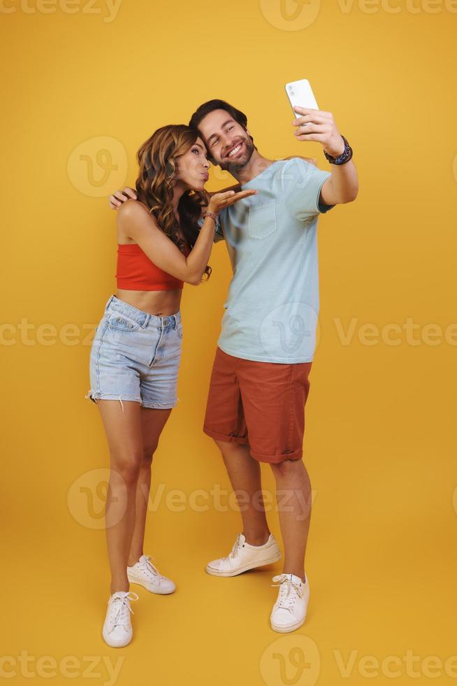 toda la longitud de una hermosa joven pareja sonriente haciéndose selfie mientras se enfrenta a un fondo amarillo foto