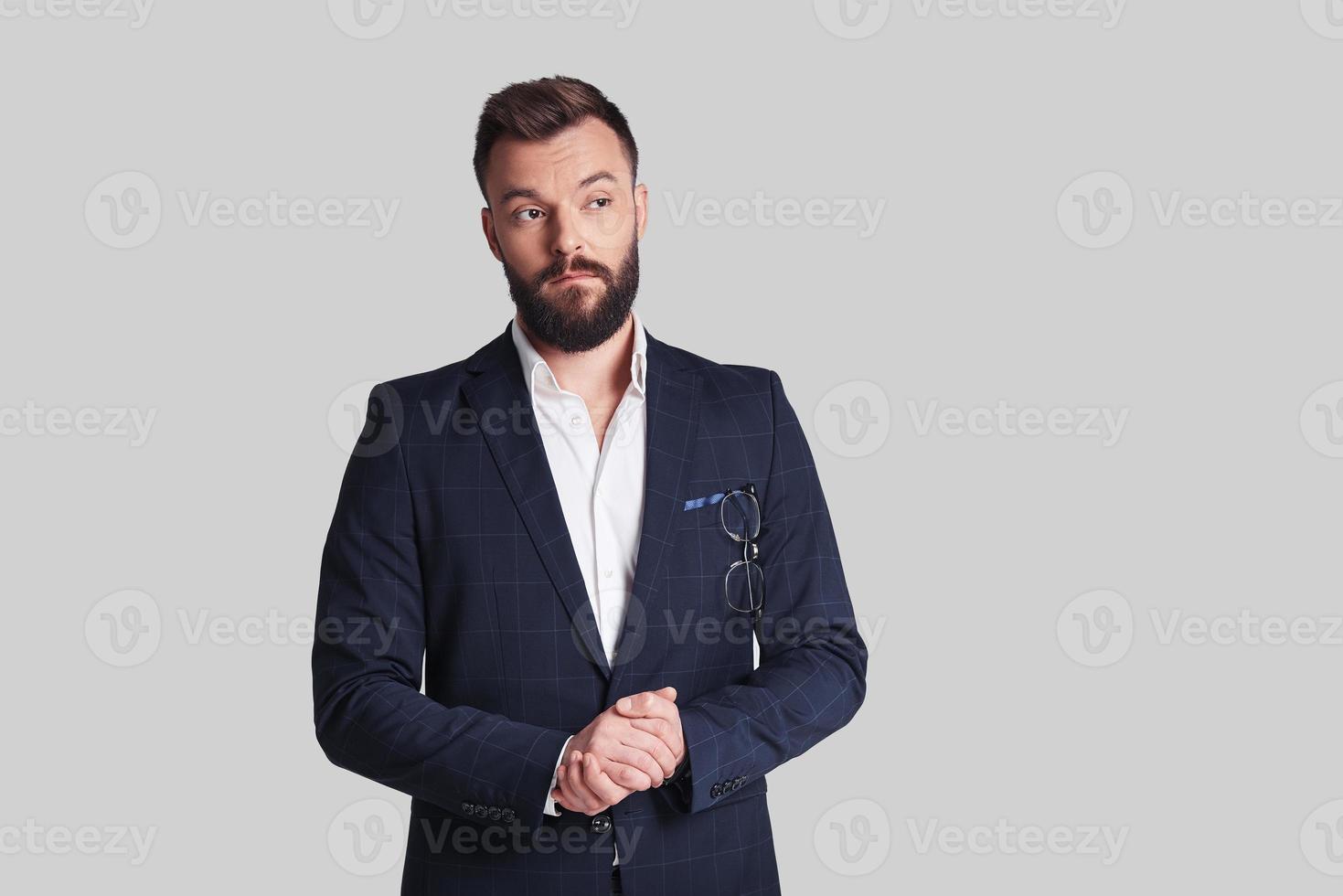 Thinking about... Handsome young man in formalwear keeping hands clasped and looking away while standing against grey background photo